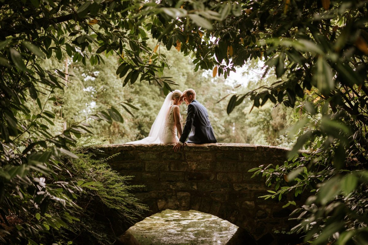 A newly married couple sharing a quiet moment on a stone bridge beneath leafy trees